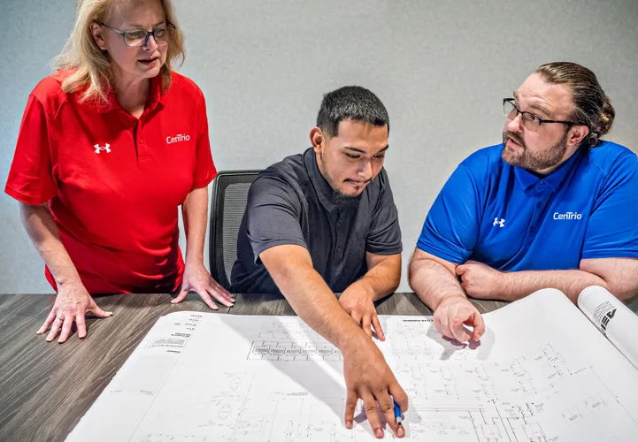Photo of a female CenTrio associate in a red shirt along with a male CenTrio Associate in a blue shirt talking with a gentleman in a black shirt at table looking at schematics.