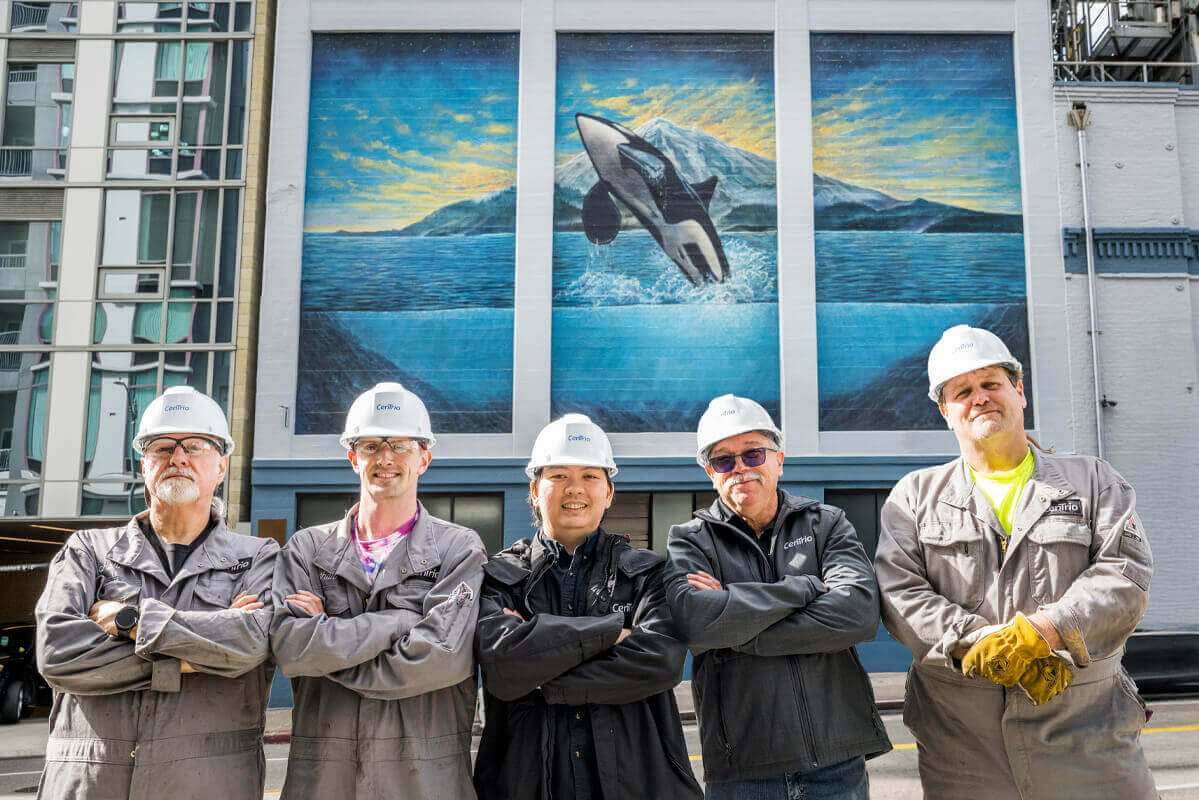 Image of five CenTrio Seattle operators standing side-by-side in front of our Seattle Plant with a mural of an orca behind them that’s painting on the wall of our plant building.