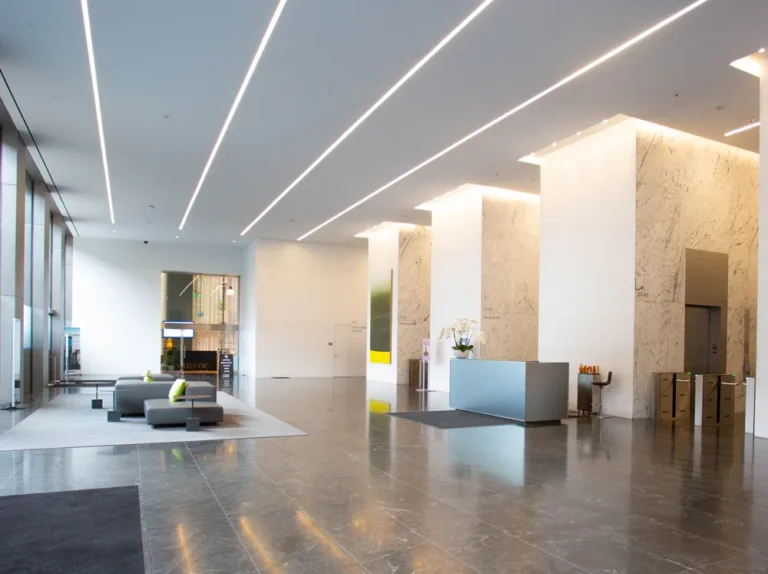 Modern office lobby with marble columns, polished floor, reception desk, and lounge seating under sleek linear ceiling lights.