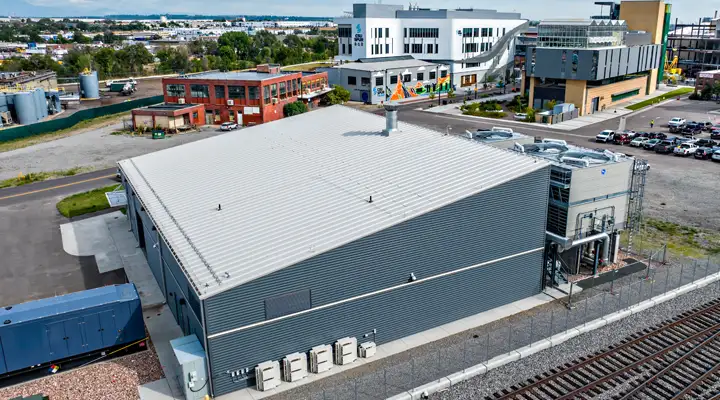 Aerial view of a modern industrial facility with a large gray metal building, ventilation systems, and adjacent railway tracks, surrounded by nearby commercial and industrial buildings.